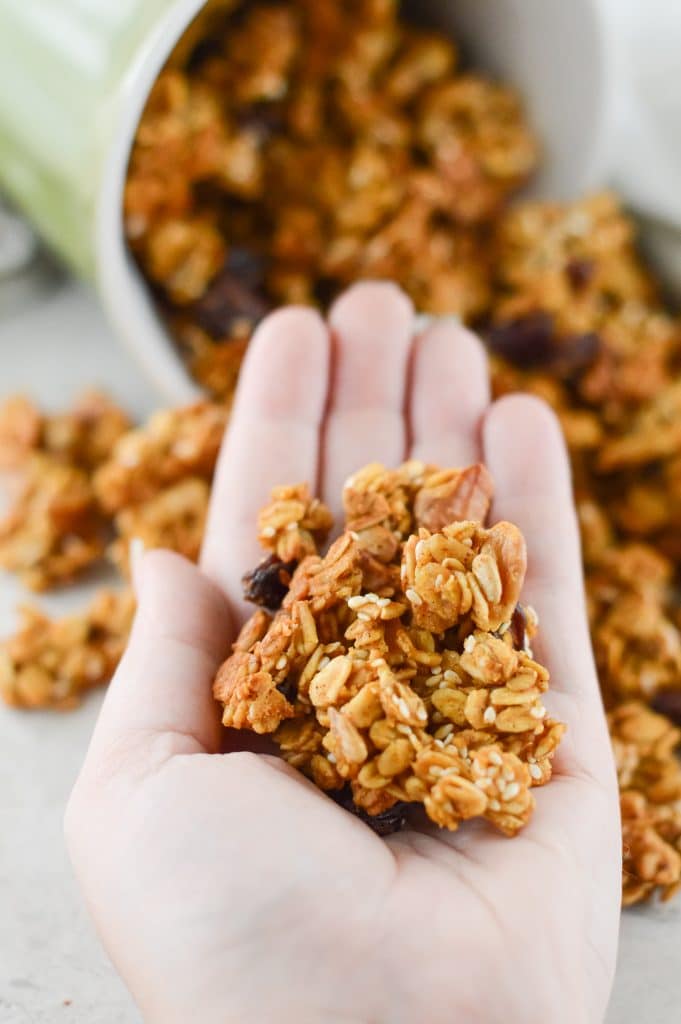 a hand holding a cluster of pumpkin spice granola with a cup of more granola spilling onto a white table in the background