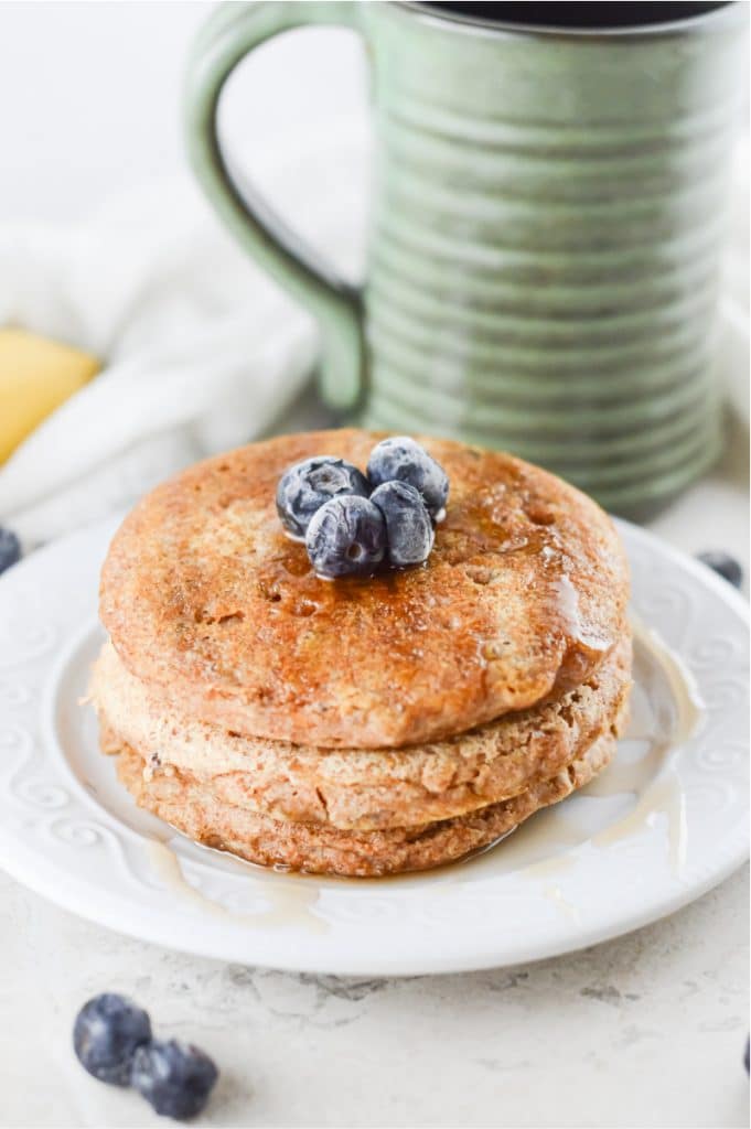 stack of vegan pancakes on a white plate, topped with maple syrup and blueberriers. Green mug in the background.