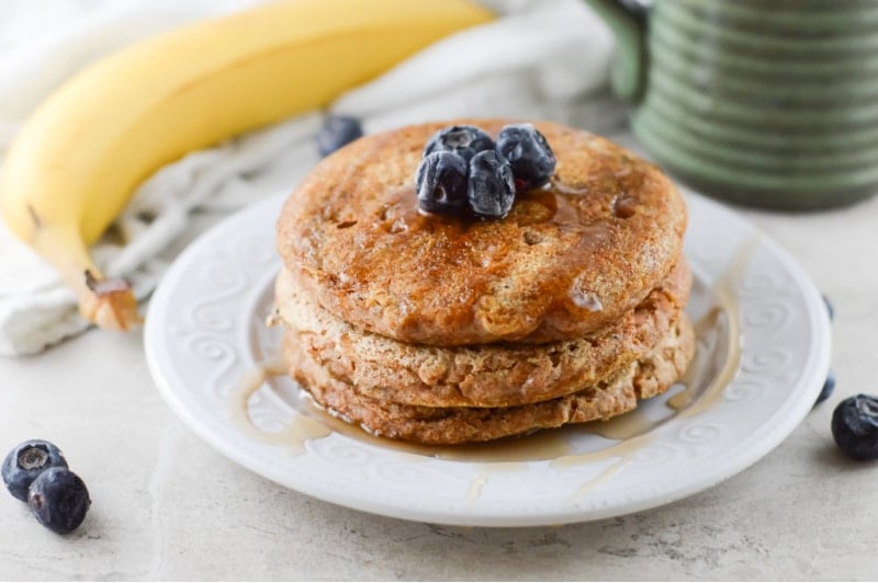 stack of pancakes on a white plate topped with maple syrup and blueberries. green mug in the background