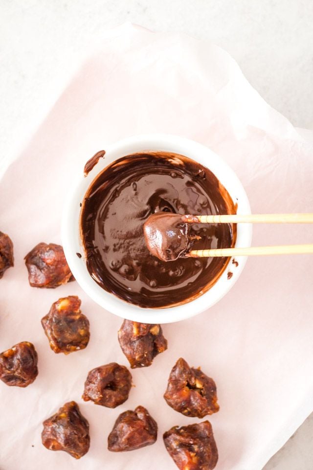 A pair of chopsticks being used to dip macadamia balls into melted chocolate