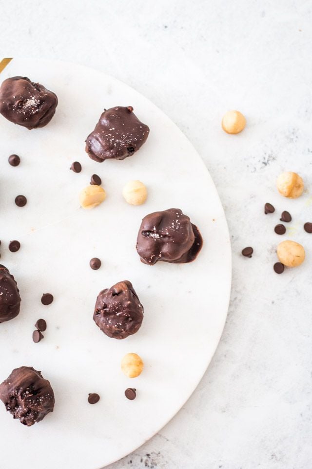 A platter of chocolate covered caramel macadamia nuts with a sprinkling of chocolate chips and macadamia nuts surrounding