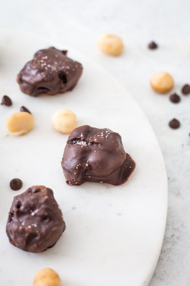 Chocolate covered macadamia nuts on a white platter surrounded by chocolate chips and whole macadamia nuts