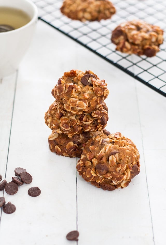 stack of crunchy oatmeal cookies with chocolate chips spread around and a cooling rack with more oatmeal cookies and a cup of tea in the background