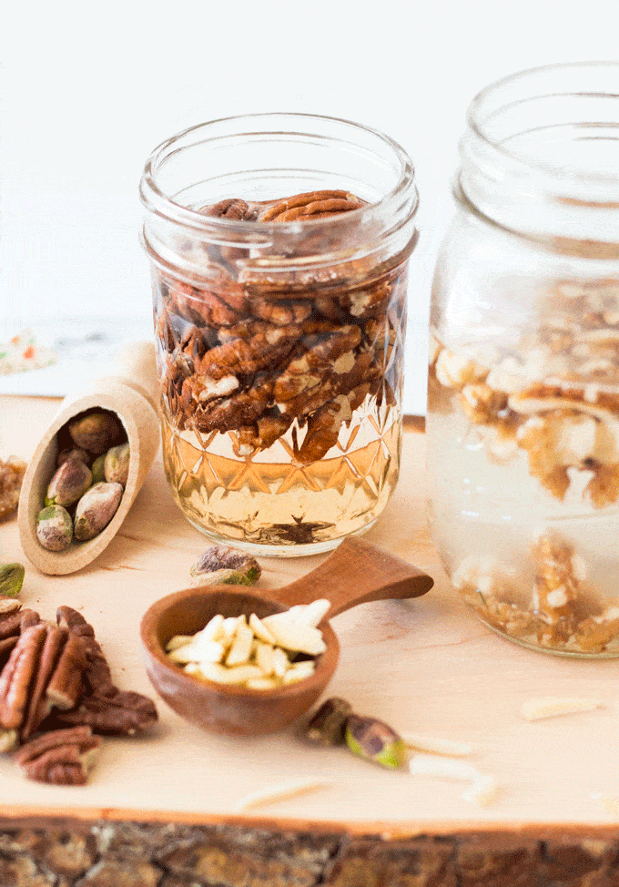 A wooden chopping board holding glass jars of nuts soaking in water and spoonfuls of various nuts used for making Homemade Nut Milk