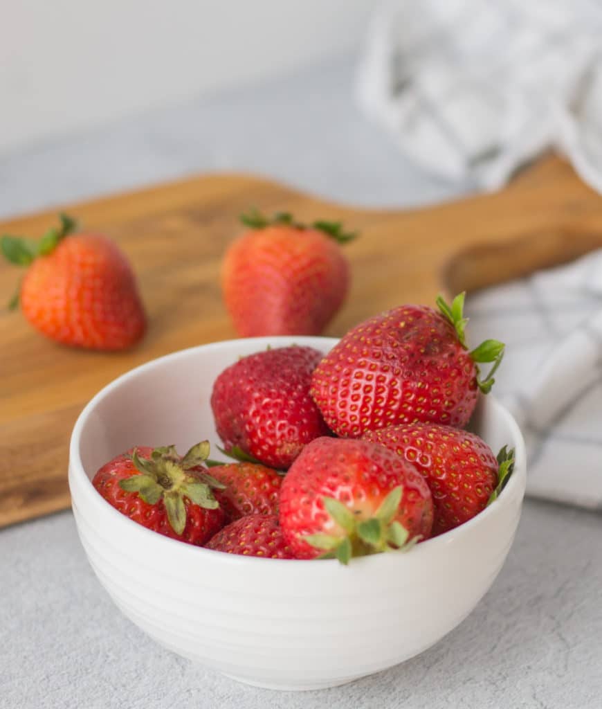 Fresh strawberries in a white bowl with a wooden chopping in the background and a couple of strawberries spread around