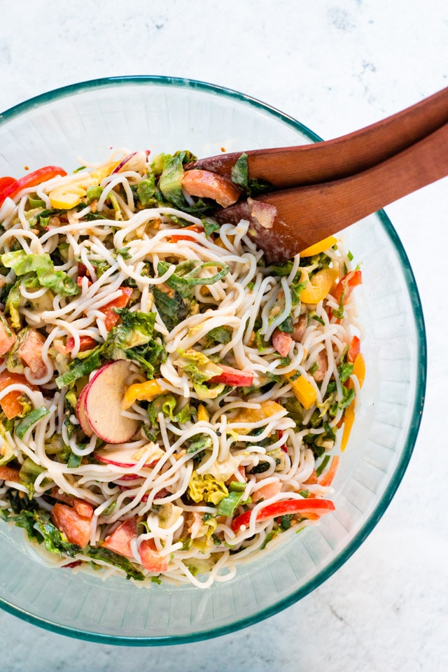 All of the ingredients for cold peanut noodle salad added together in a large glass bowl and tossed with wooden salad servers
