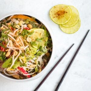 Cold peanut noodle salad served in a large white bowl against a grey background with wooden chopsticks and chilli covered lime in the background