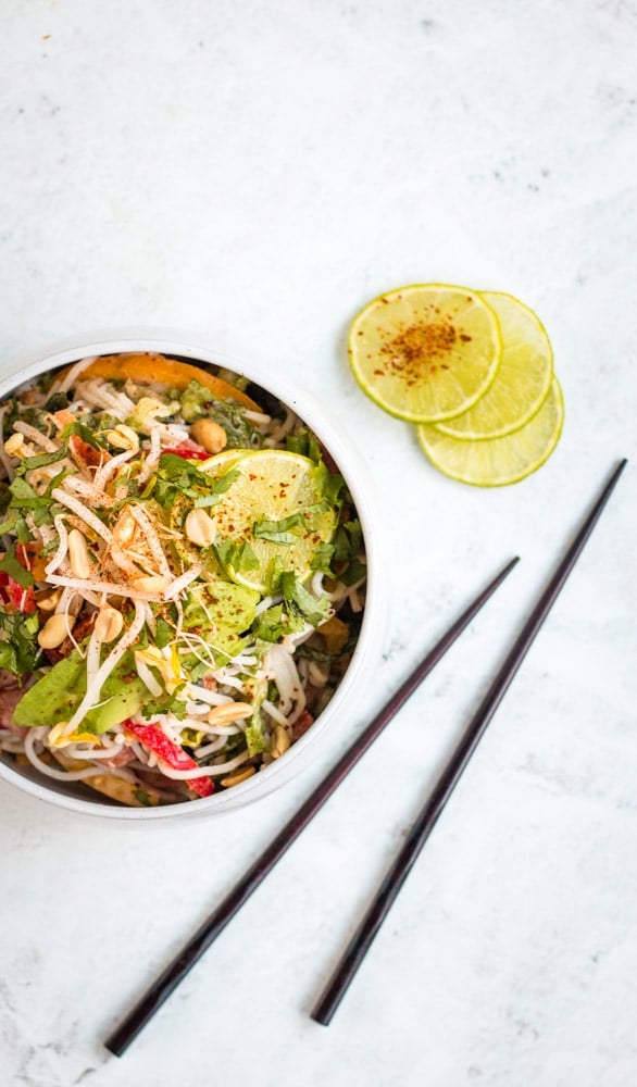 Cold peanut noodle salad served in a large white bowl against a grey background with wooden chopsticks and chilli covered lime in the background