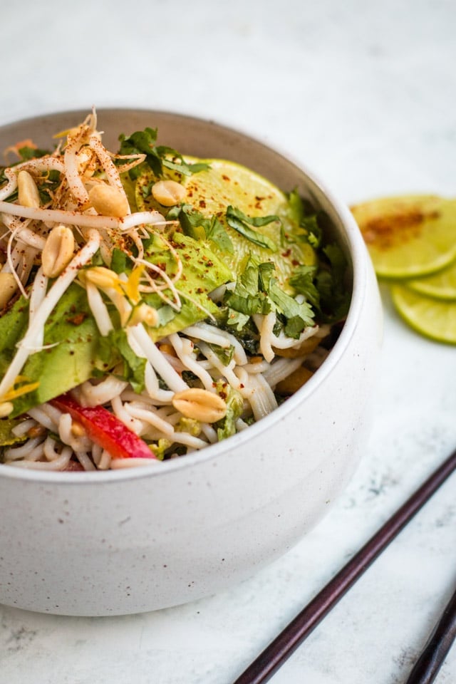 Cold peanut noodle salad served in a large white bowl against a grey background with wooden chopsticks and chilli covered lime in the background