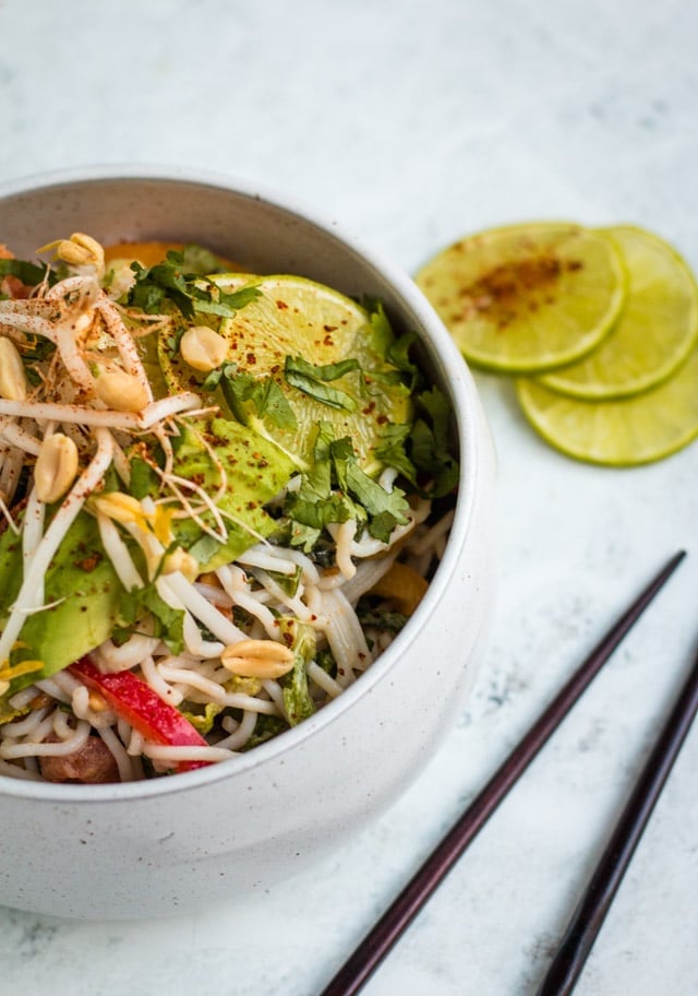 Cold peanut noodle salad served in a large white bowl against a grey background with wooden chopsticks and chilli covered lime in the background