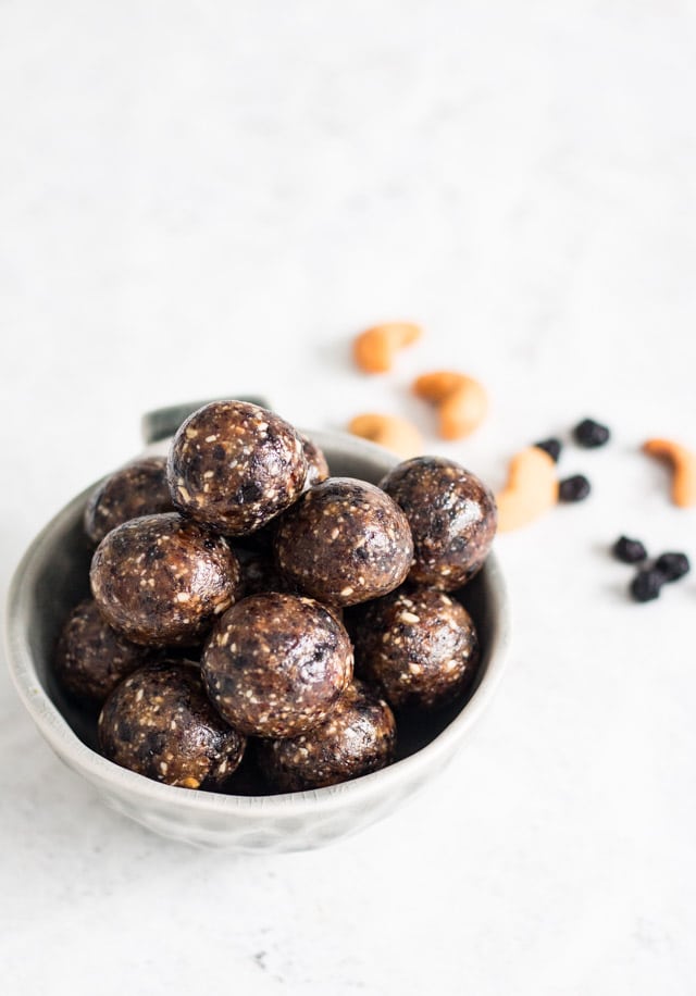 Blueberry bliss balls served in a small grey bowl against a white background with sprinkles of cashews and dried blueberries in the background