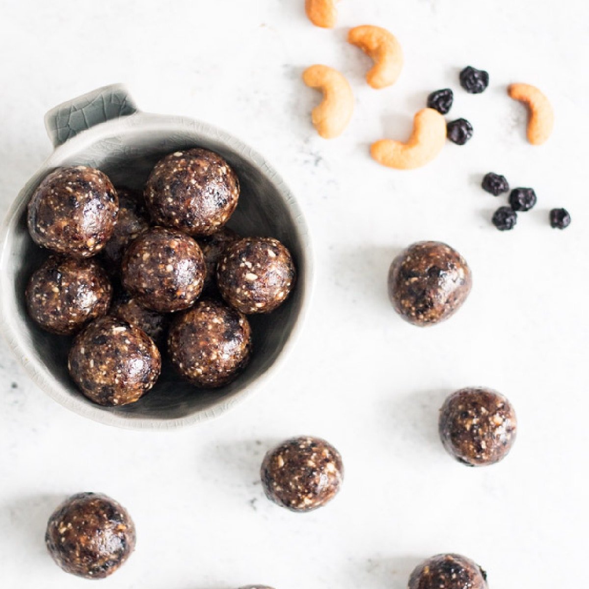 Overhead shot of cashew bliss balls arranged against a white background with a sprinkling of cashews and dried blueberries in the background
