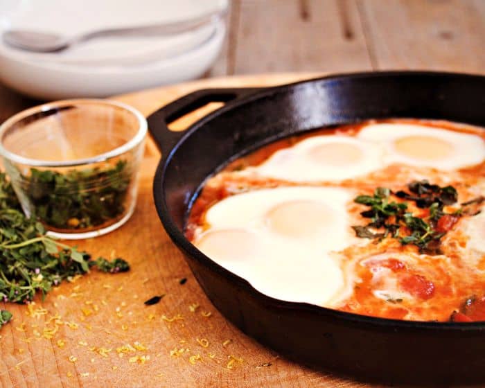 slow steamed eggs on top of a tomato based sauce in a cast iron saucepan with fresh herbs sprinkled around the chopping board