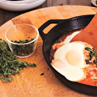 slow steamed eggs on top of a tomato based sauce in a cast iron saucepan with fresh herbs sprinkled around the chopping board