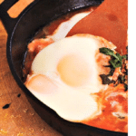 slow steamed eggs on top of a tomato based sauce in a cast iron saucepan with fresh herbs sprinkled around the chopping board