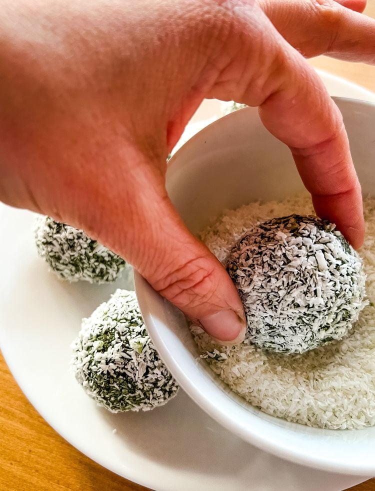 A hand rolling green bliss balls in a small bowl of desiccated coconut