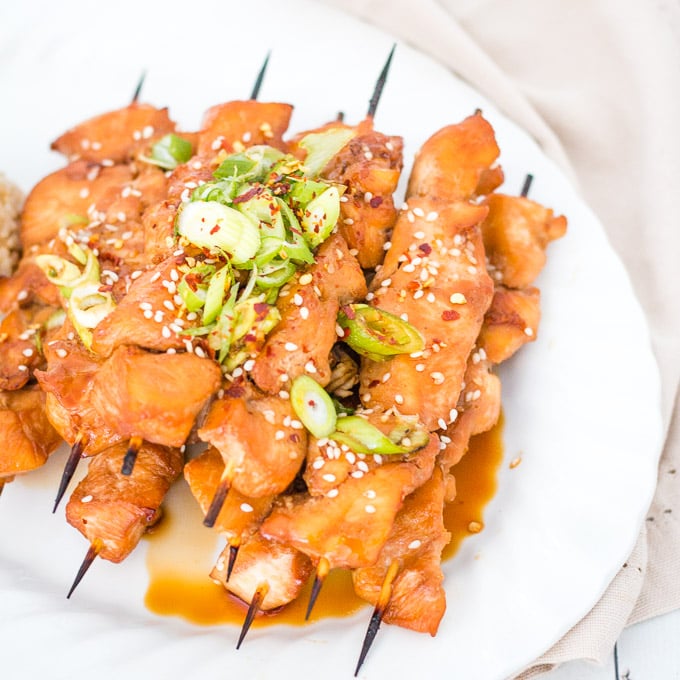 pile of teriyaki chicken skewers on a white plate topped with chopped spring onions