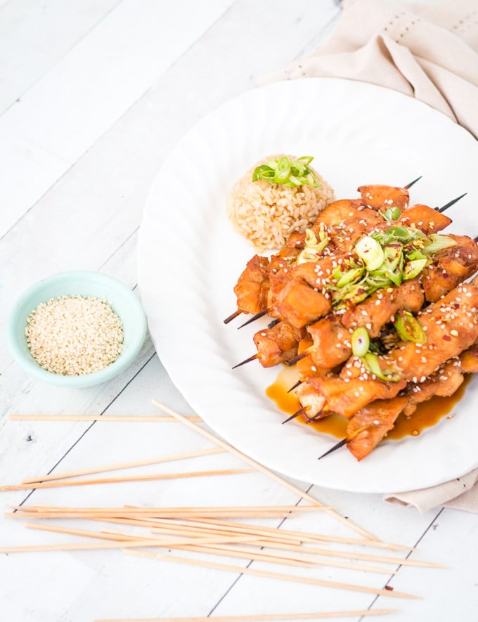 Teriyaki chicken skewers piled onto a white plate with a green bowl of sesame seeds on the side