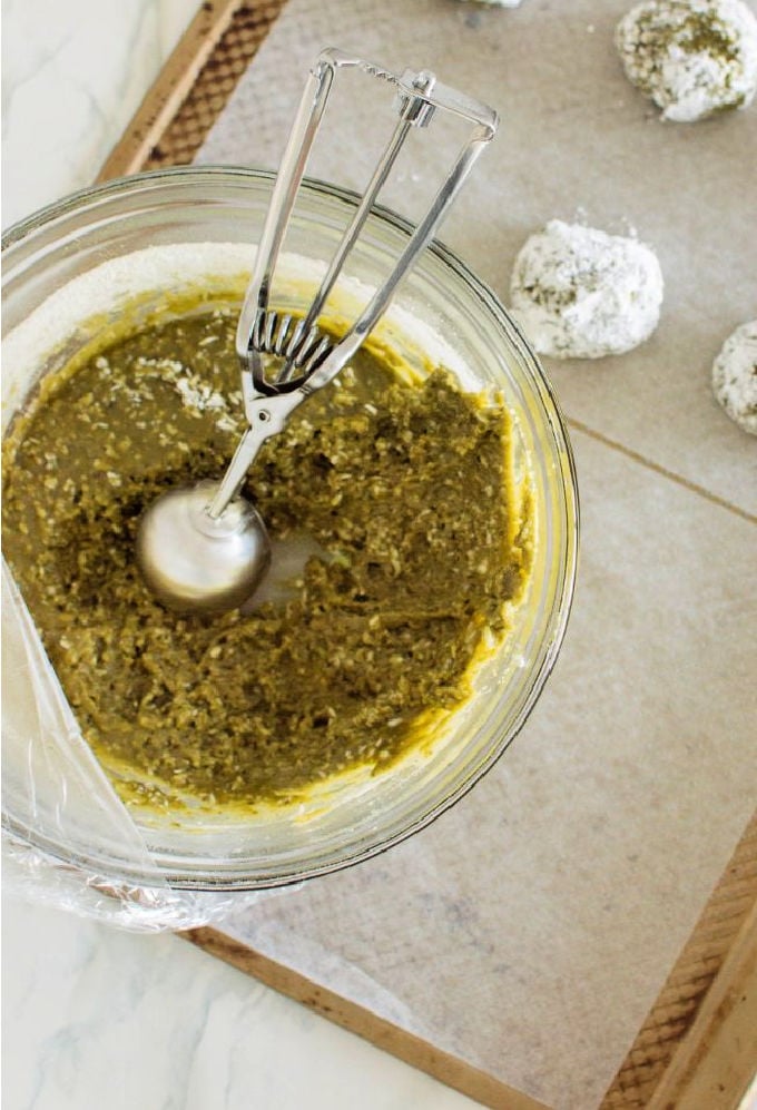 Large glass bowl with green energy ball mixture and some energy balls in the background that have been rolled in coconut