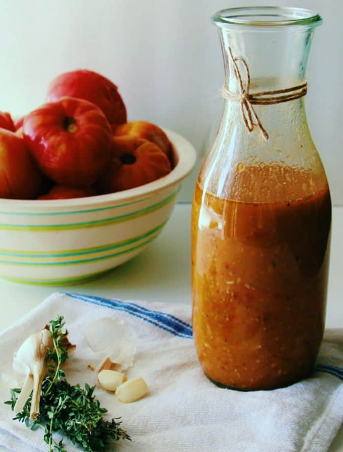 homemade heirloom tomato sauce in a large glass just with a bowl of fresh tomatoes in the background, and a bunch of fresh herbs on the side