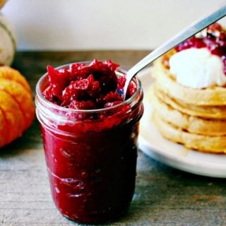 Honey Sweetened Vanilla and Fresh Cranberry Butter served in a glass jar with a pile of waffles in the background topped with the spread.