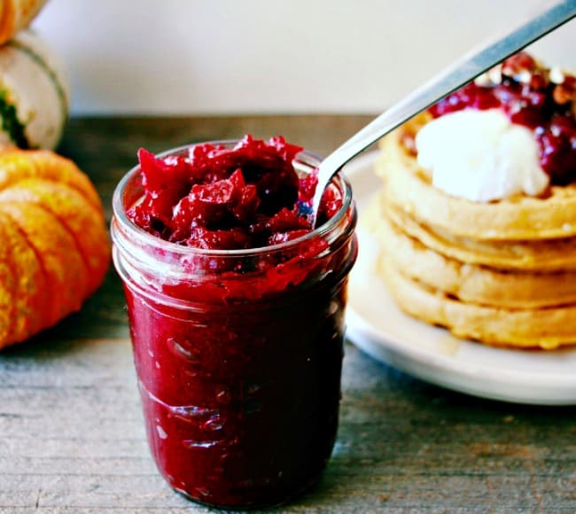 Honey Sweetened Vanilla and Fresh Cranberry Butter served in a glass jar with a pile of waffles in the background topped with the spread.