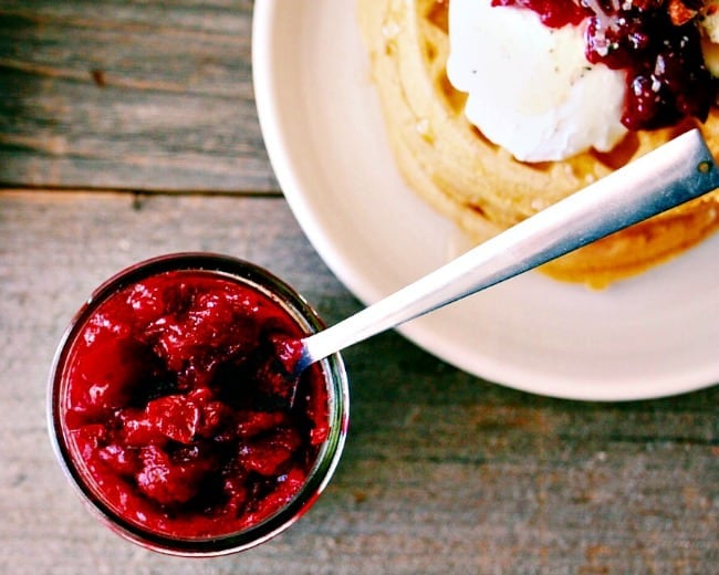 Overhead shot of Honey Sweetened Vanilla and Fresh Cranberry Butter served in a glass jar with a pile of waffles in the background topped with the spread.