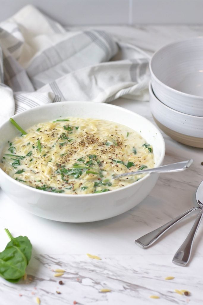 Creamy orzo with spinach served in a white bowl with some fresh spinach and sprinkles of uncooked orzo in the foreground