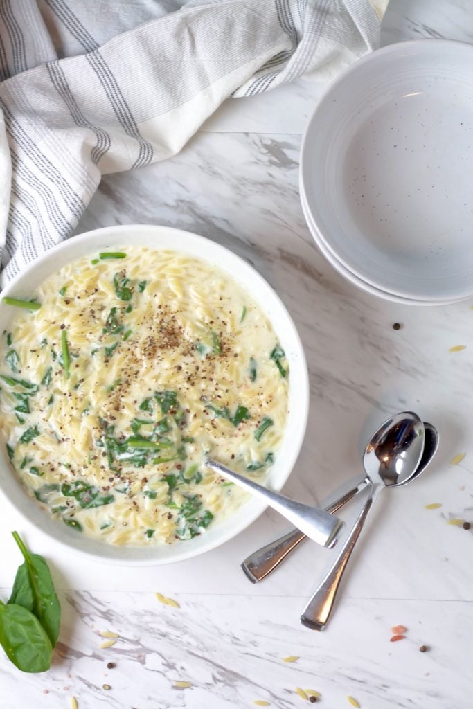 Overhead shot of creamy orzo with spinach served in a white bowl with some fresh spinach and sprinkles of uncooked orzo in the foreground