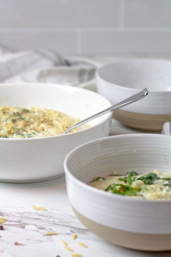 Serving shot of creamy orzo spinach in a large white and tan bowl being served up into white bowls by a serving spoon