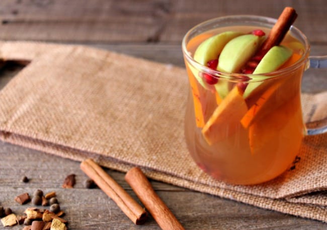 Cider served in a glass mug with a cinnamon stick, apple slice and some pomegranate. There are mulling spices and cinnamon sticks surrounding the mug