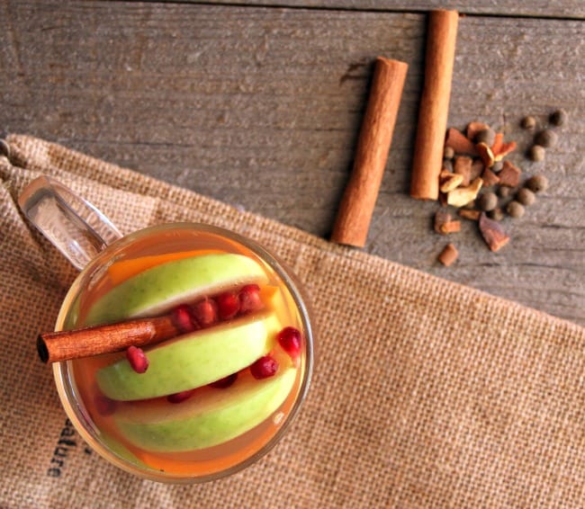 overhead shot of an apple cider served in a glass mug with apple slice, pomegranate arils, and a cinnamon stick. There are cinnamon sticks and mulling spices on the side