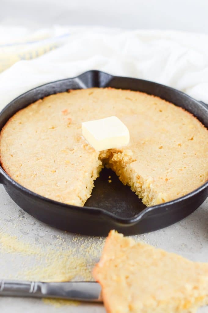 a wedge of cornbread being served from a cast iron skillet containing some homemade corn bread with a dollop of honey butter  on top
