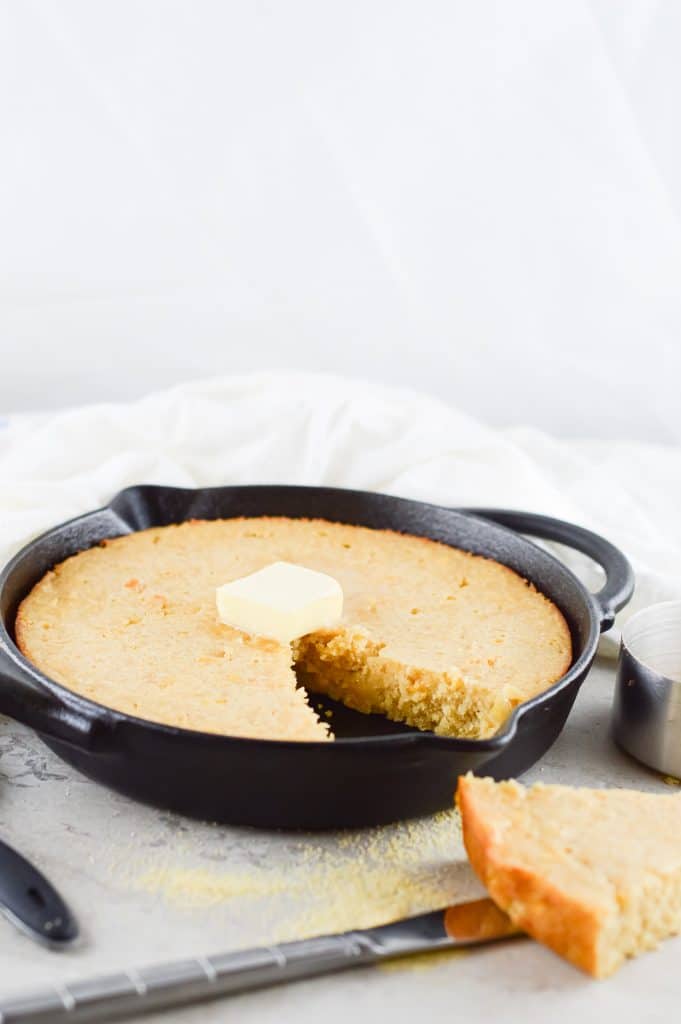 Homemade honey butter cornbread in a cast iron skillet with a dollop of butter on top and a slice removed and sitting on the side