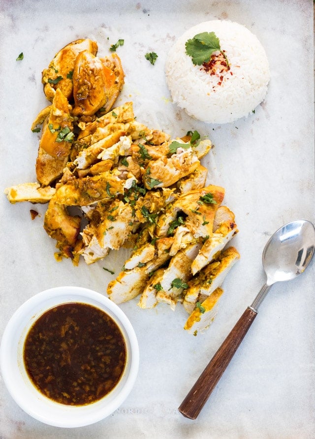 overhead shot of turmeric baked chicken sliced up with a bowl of sauce on the side, a mound of rice and a wooden handled spoon for serving