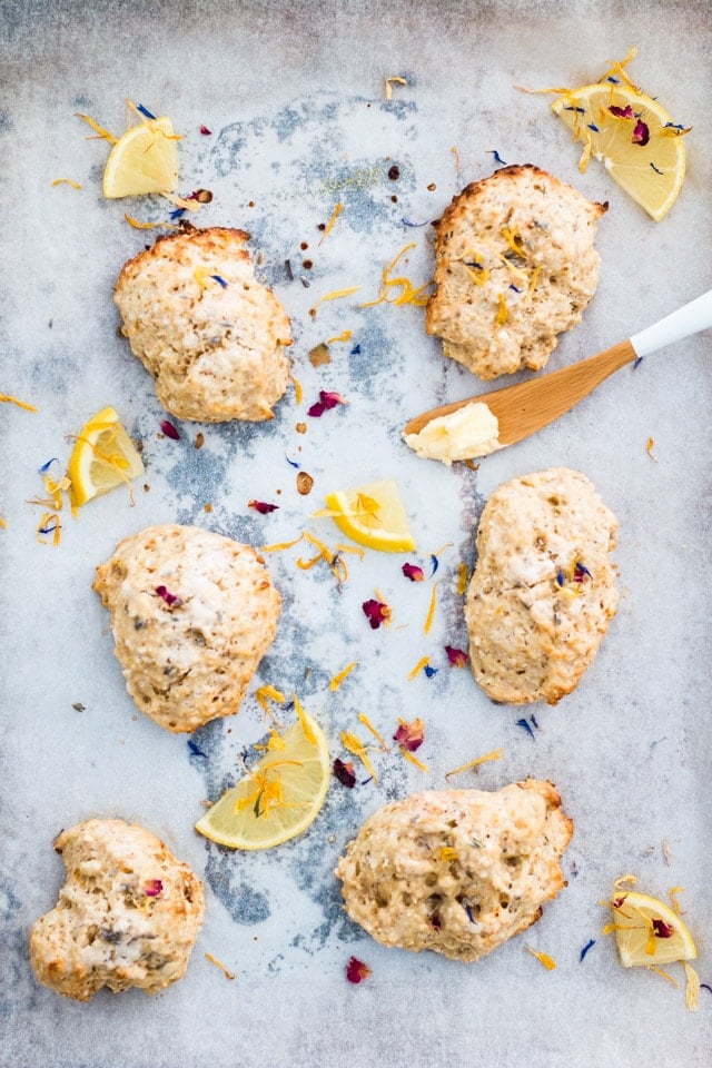 Overhead shot of 6 lemon and lavender scones on a baking tray and sprinkled with edible flowers and small lemon wedges with a butter knife on the side