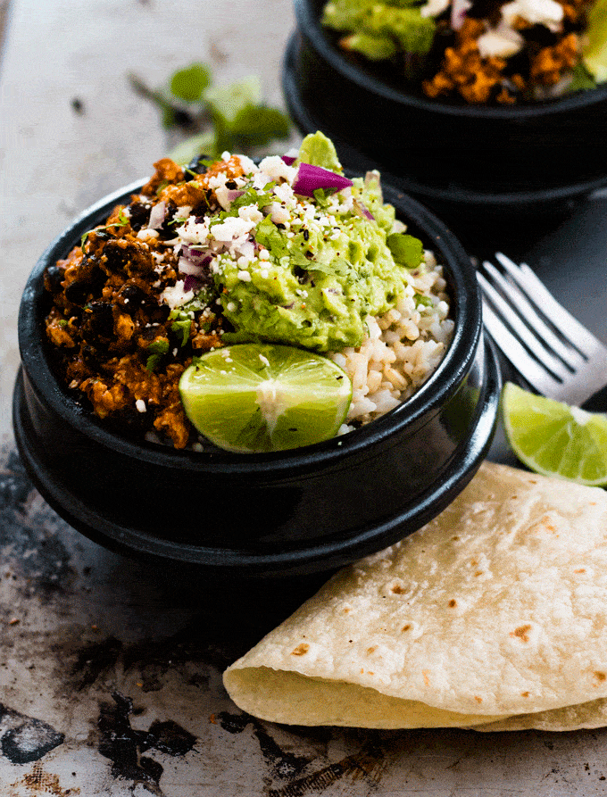 Up close shot of a black bowl containing chipotle tofu and brown rice topped with guacamole, red onion and lime wedges. There is also a tortilla on the side.