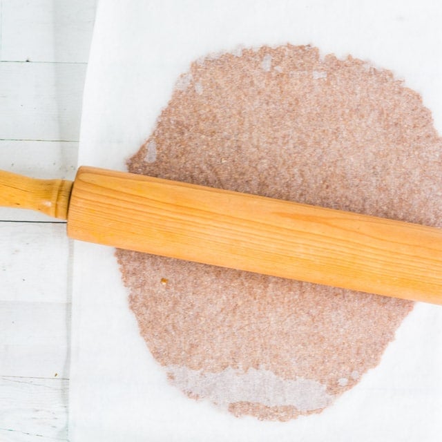 raw graham cracker dough between two pieces of parchement paper, being rolled with a wooden rolling pin