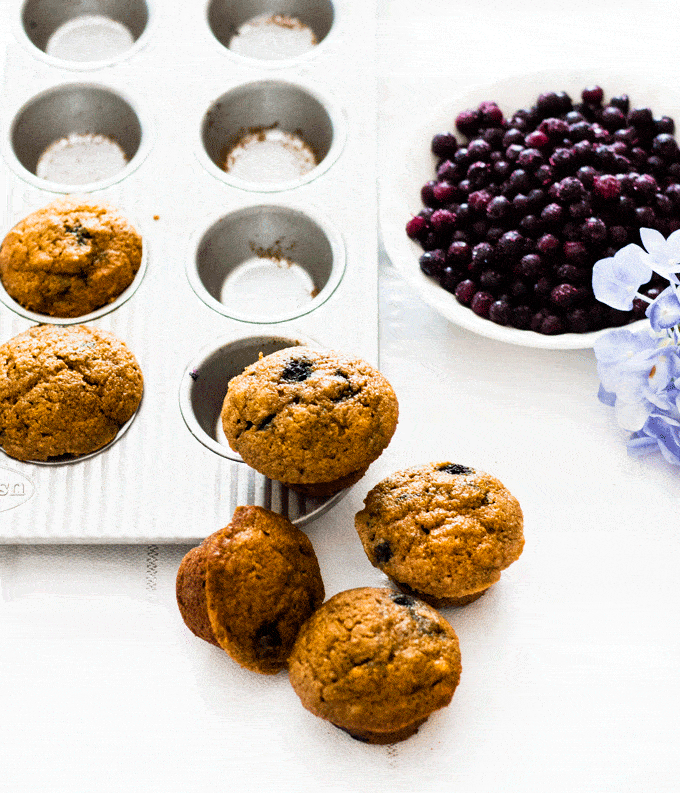 A pile of mini muffins resting on a muffin tin with a bowl of blueberries in the background
