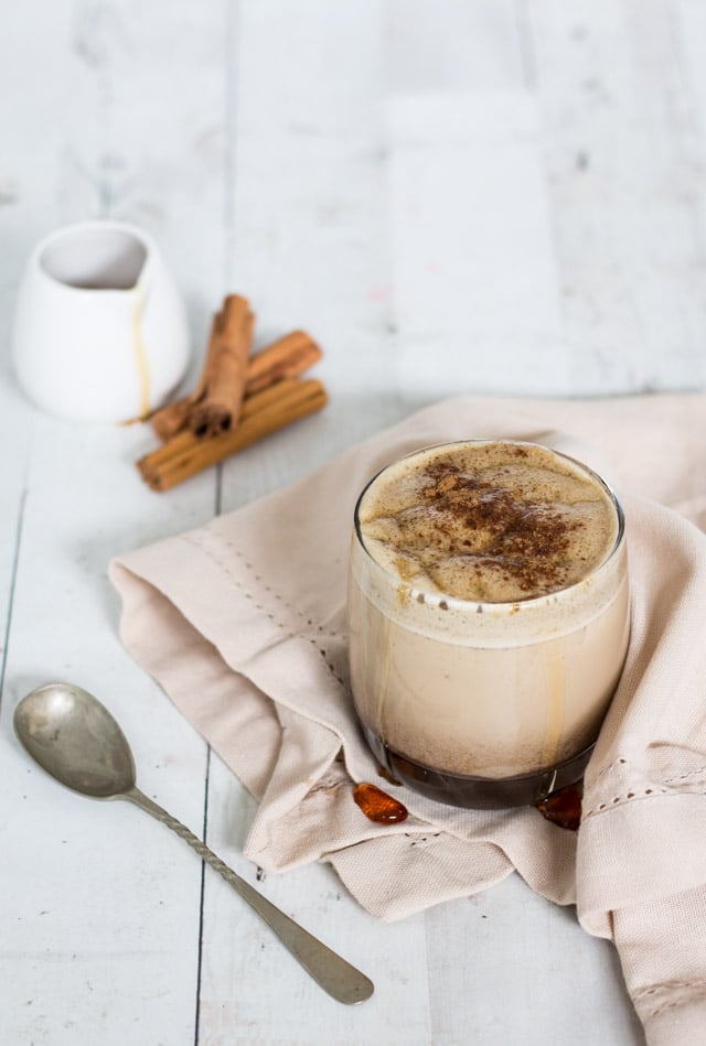 Green tea latte with maple syrup dripping down the side of the glass and a small jug of maple syrup and cinnamon sticks in the backgroun