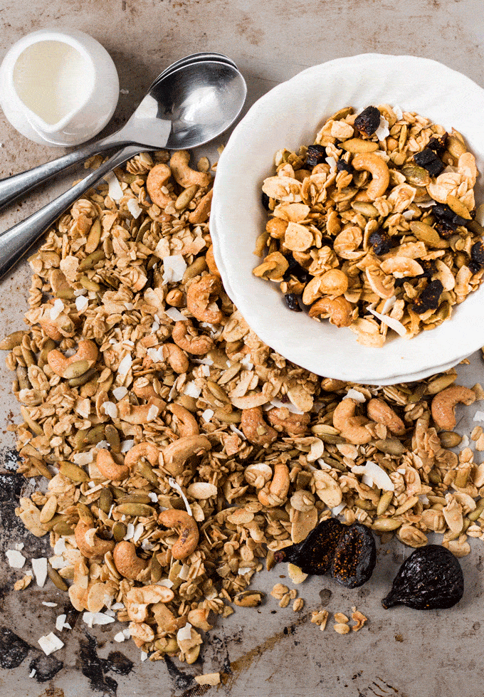 Homemade granola served in a white bowl with a side of milk and dried figs. The granola is surrounded by loose freshly baked granola on a metal baking sheet.