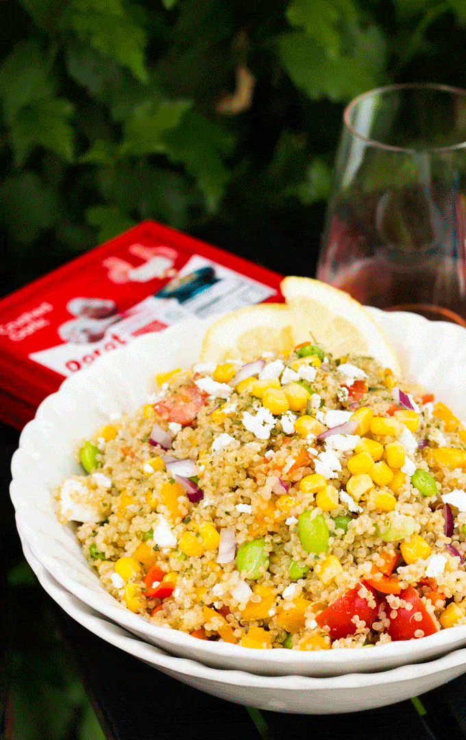 A large white bowl containing a quinoa and chopped vegetable cold salad with a glass of water in the background