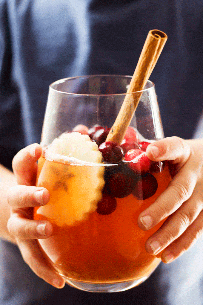 a childs hands holding forth a glass of hot cranberry apple cider in a wine glass with floating cranberries, a crimped apple slice and a large cinnamon stick poking out of the glass