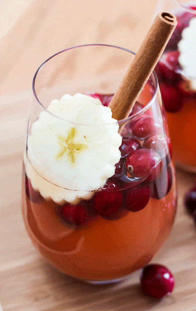 wine glass containing hot cranberry apple cider with cranberries, a crimped apple slice and a long cinnamon stick 