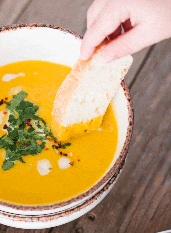 Ginger carrot and turmeric soup in an off-white bowl, topped with cilantro and with some bread being dipped in