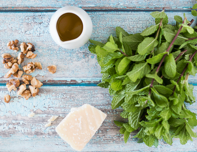 all the ingredients for Mint Pesto laying on a blue background. mint, parmesan, oil and walnuts