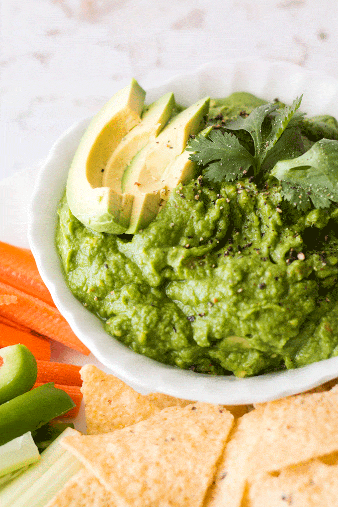 Green pea Guacamole in a white bowl surrounded by crudités.