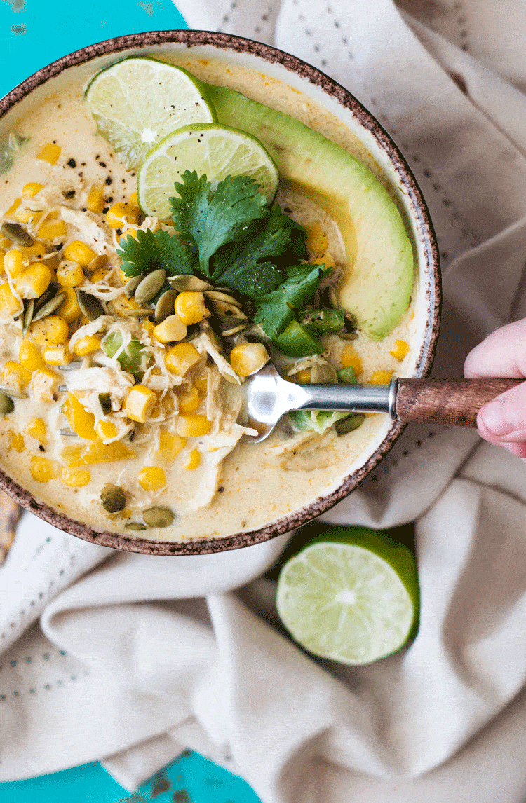 A spoon being dipped into a large bowl of white chili topped with avocado and cilantro