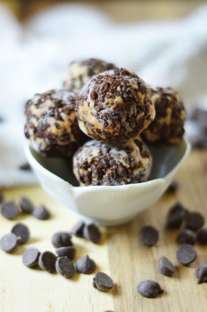 A pile of Chocolate Cheesecake Fat Bombs in a white bowl on a wooden background surrounded by chocolate chips