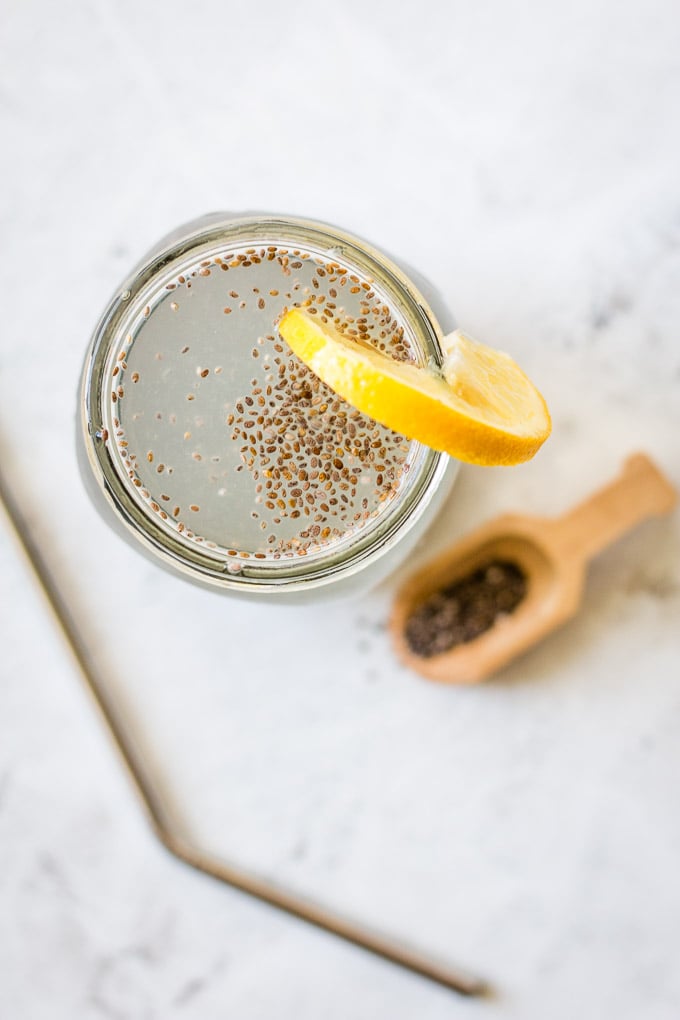 top view of chia water in a glass with chia seeds floating on top, a slice of lemon, and a small spoon full of seeds on the side